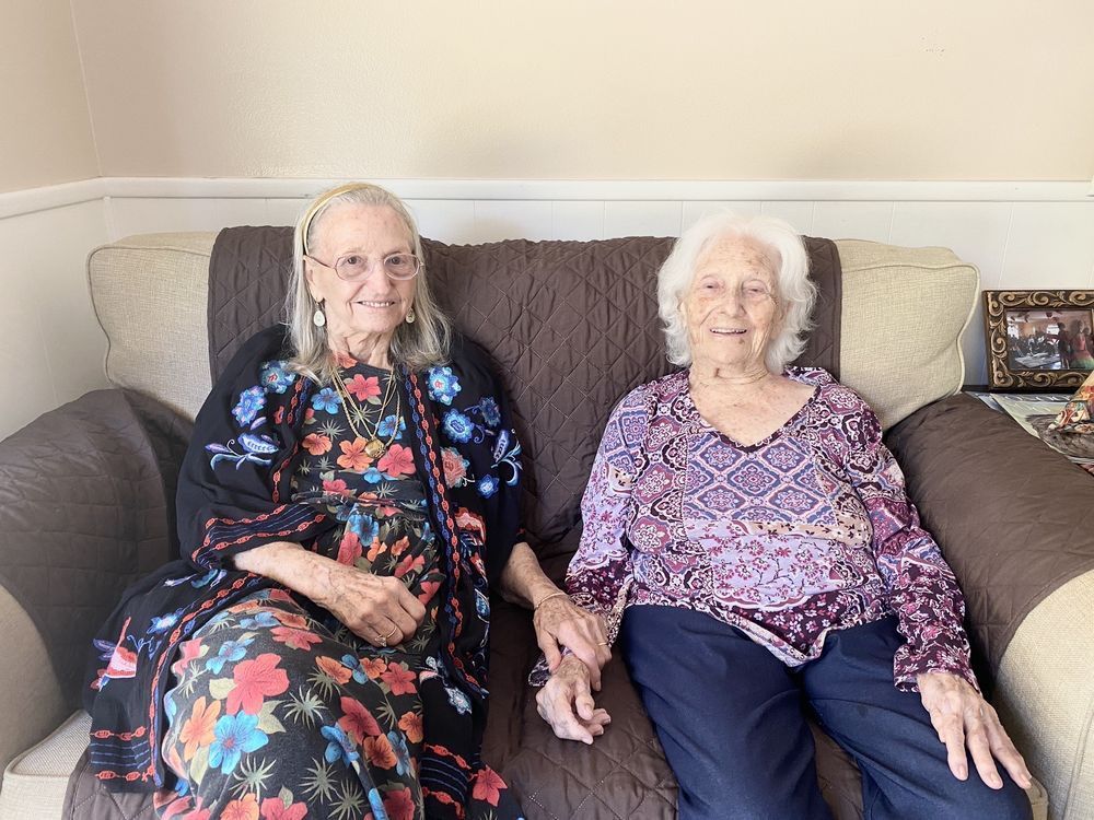 Two older women are sitting on a couch holding hands.