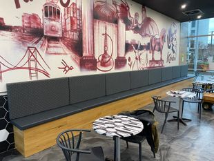 Restaurant interior with a long upholstered bench against a mural, small tables, and chairs.