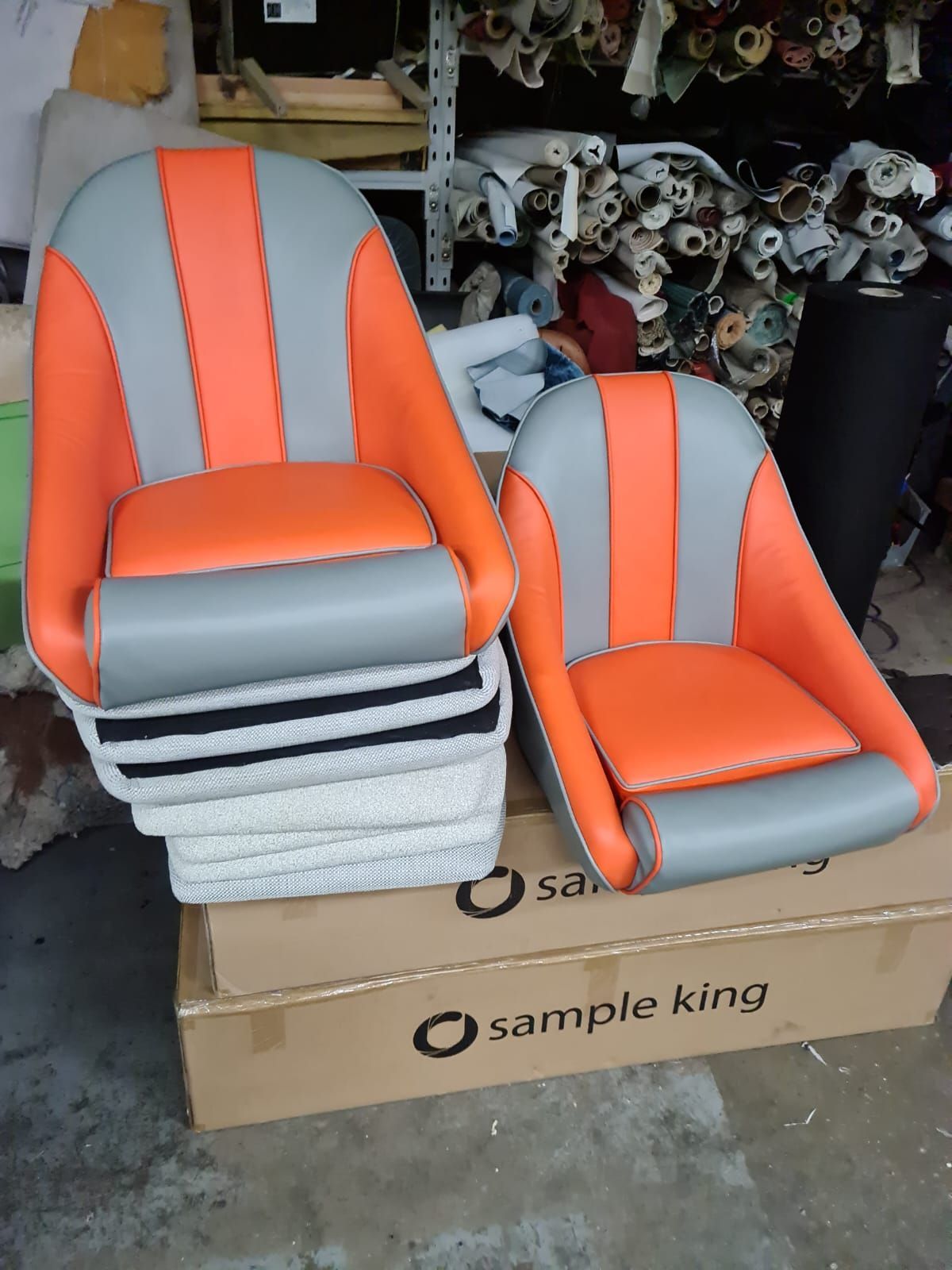 Two boat seats, orange and gray, displayed on boxes with rolls of fabric in the background.