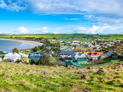 A scenic coastal town with houses, green fields, and a beach under a blue sky with clouds.