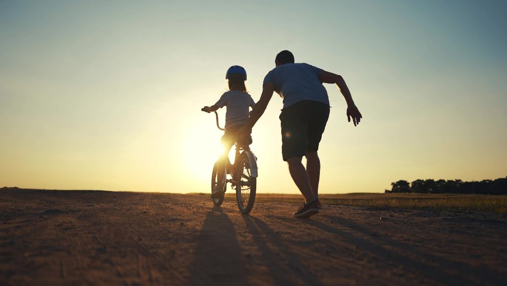 Father helping child learn to ride a bike at sunset on a dirt road. Silhouette of figures against the bright sun.
