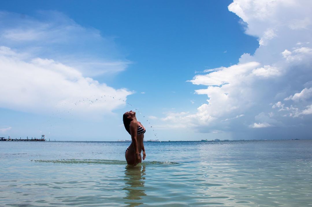 Woman in a bikini in shallow water, looking up with arms raised, water droplets in the air; blue sky with clouds.