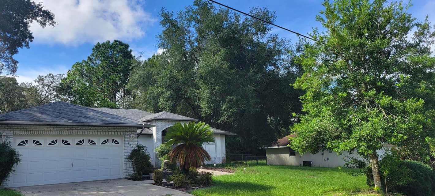 A house with a white garage door, surrounded by green trees and grass, under a blue sky with clouds.
