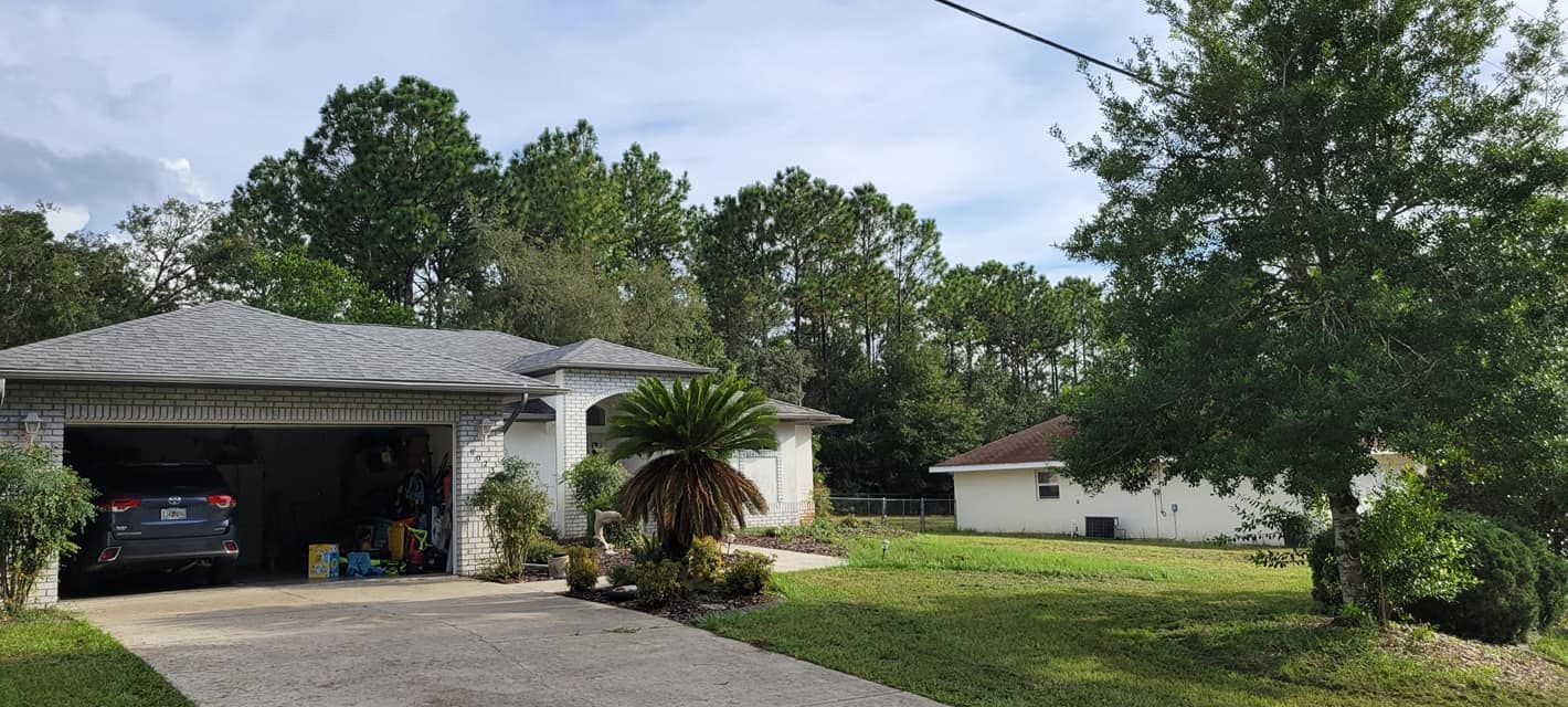 A white house with a gray roof and garage. A car is parked in the garage, trees surround the house.