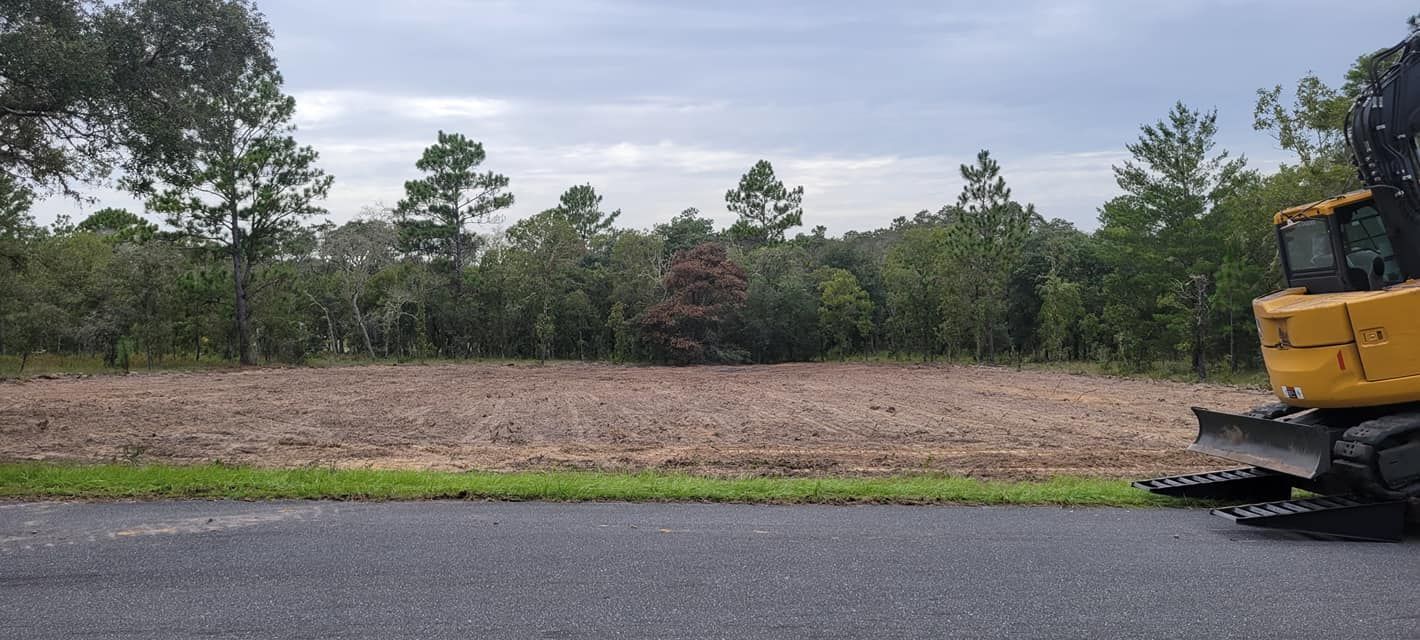 A yellow excavator on asphalt with a cleared field and tree line under a cloudy sky.