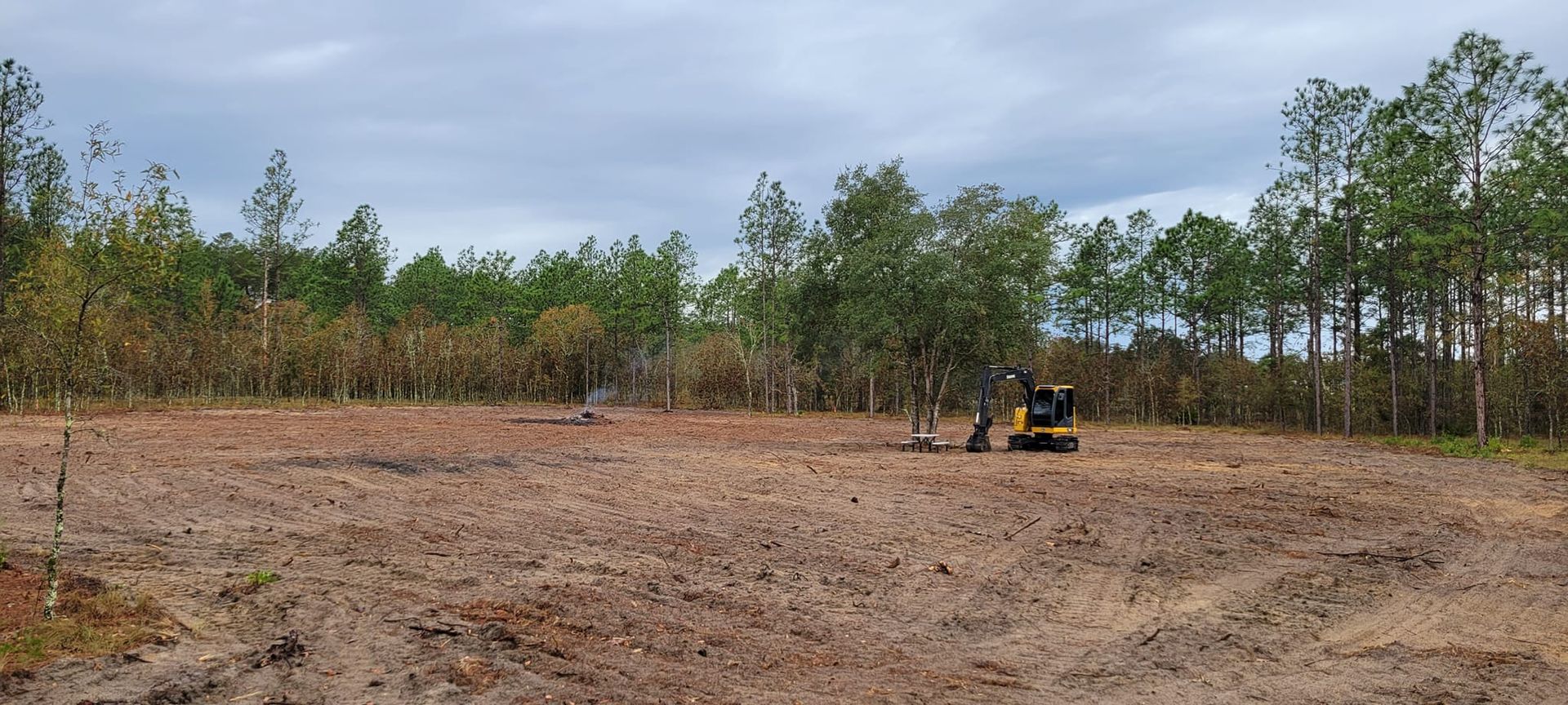 Excavator clearing a field of dirt, trees in the background, cloudy sky.
