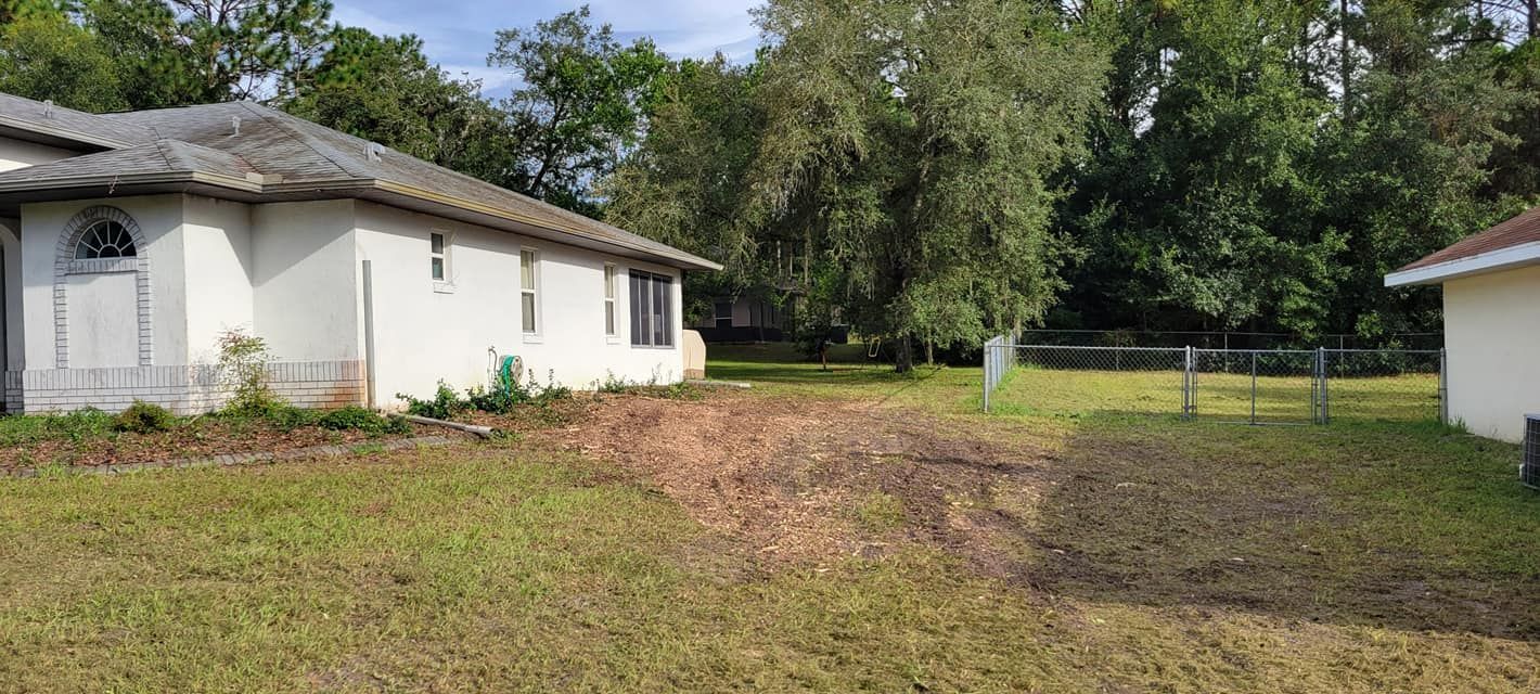 A one-story white house with a gray roof sits in a yard with a pile of mulch and trees in the background.