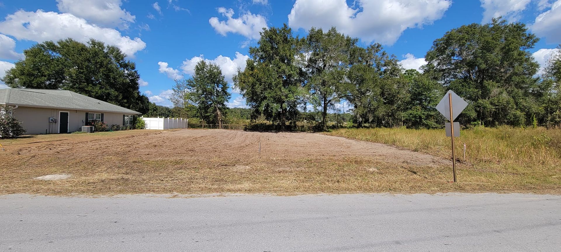 View of a vacant lot with a house on the left, trees in the background, and a road sign on the right. Blue sky.