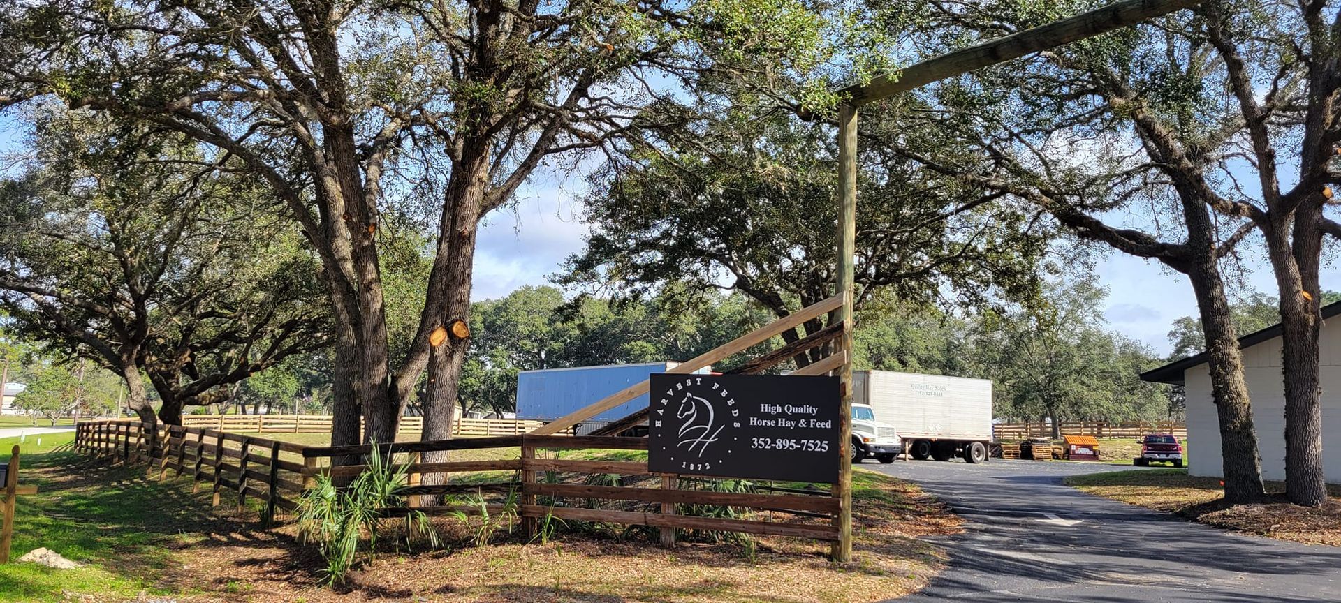 Entry to a property marked by a sign, with trees, a fence, and a distant building.