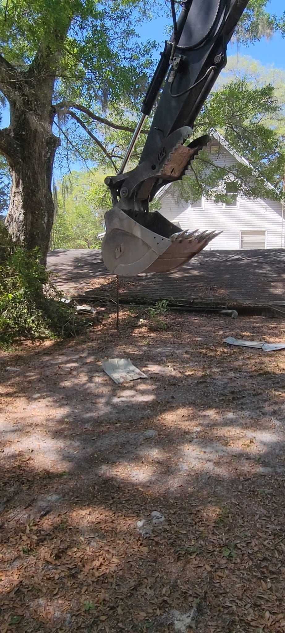 An excavator lifts its bucket in a tree-filled outdoor setting.