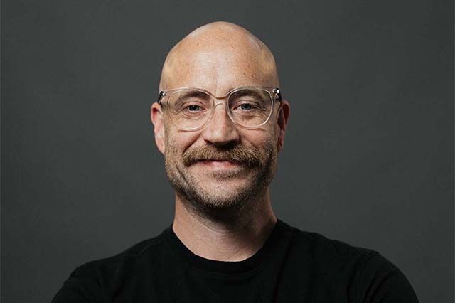 Bald man with glasses and a beard smiles outdoors, wearing a blue dotted shirt.