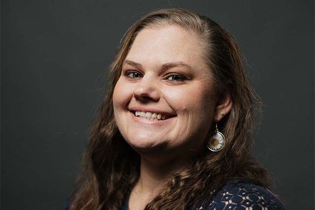 Woman with a long braid, wearing a blue shirt, smiling in front of a brick building.