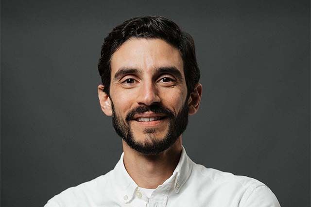 Man with glasses smiles at the camera, wearing a white button-down shirt in front of a building.