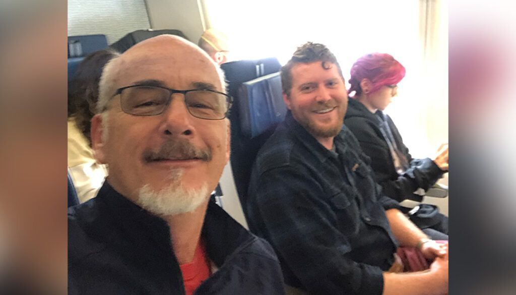 Man with glasses takes selfie with two others on a plane.