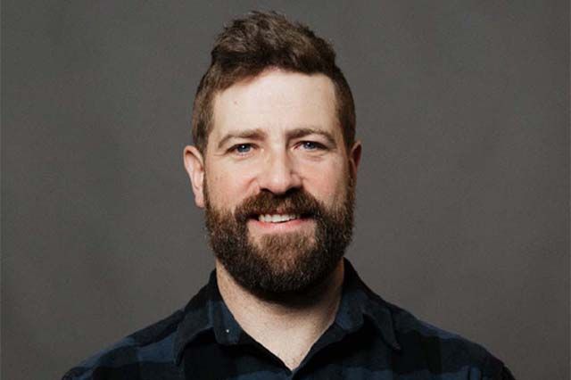 Man with beard, blue shirt, smiles outdoors. Red brick building in the blurry background.