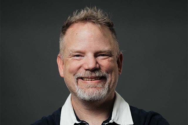Man in blue shirt smiles, standing outside a brick building.