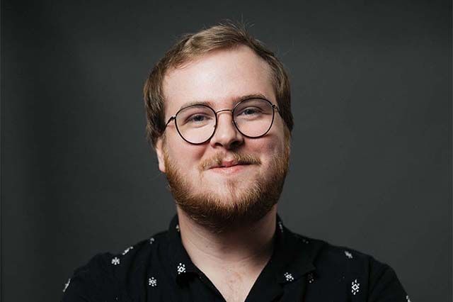 Man with glasses and fair hair smiles by a brick wall. He wears a black jacket and white shirt.
