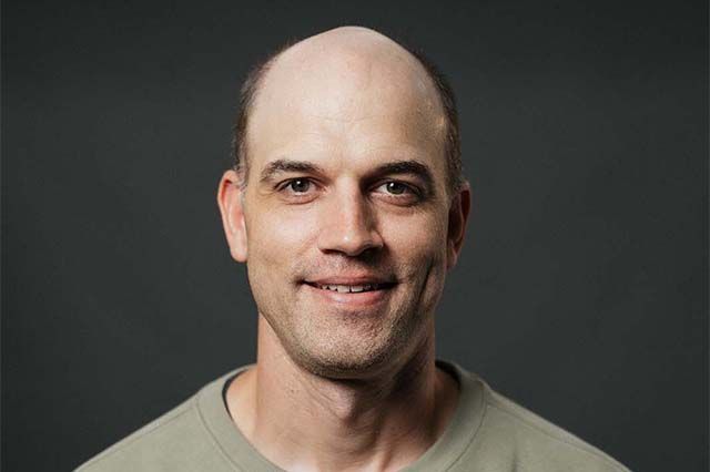 Man with a shaved head and a gray collared shirt smiles in front of a building.