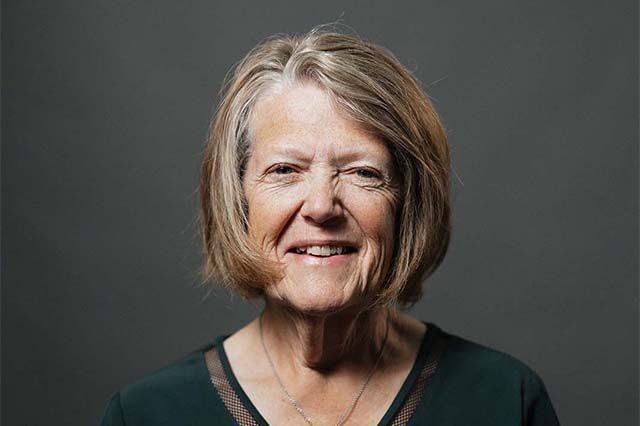 Woman with short brown hair smiling, wearing a light blue sweater, with a house in the blurred background.