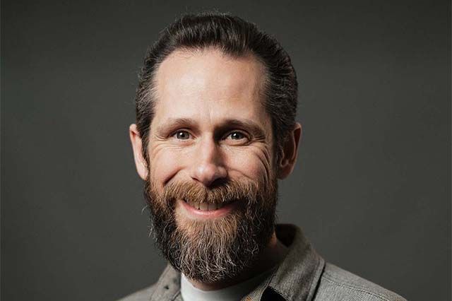 Man with dark hair and beard smiles, wearing a blue shirt outdoors, in front of a building.
