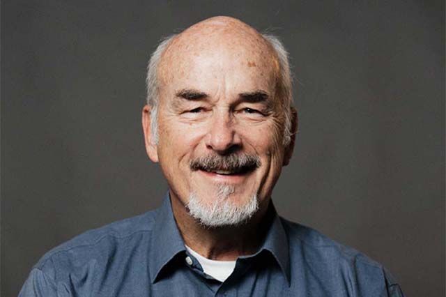 Smiling man with a beard, in a blue shirt, stands outside a building with white and brick walls.