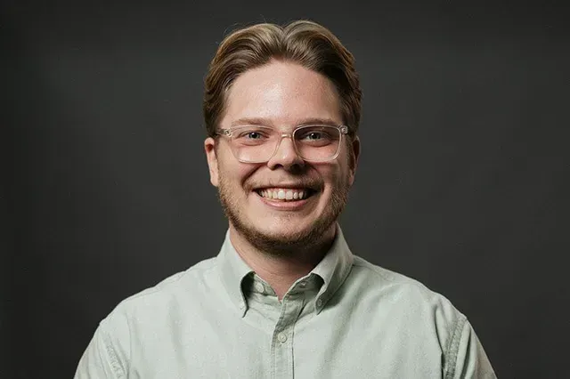 Man with glasses smiles, wearing a light green button-down shirt. Dark gray background.