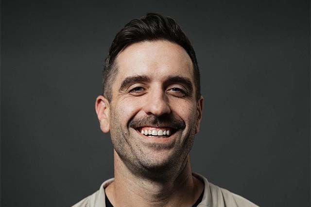 Man smiles, outdoors, wearing a black jacket over a white shirt. Green foliage background.