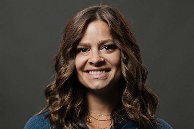 Woman with shoulder-length brown hair, smiling, wearing a black top, small necklace, outdoors with green foliage.