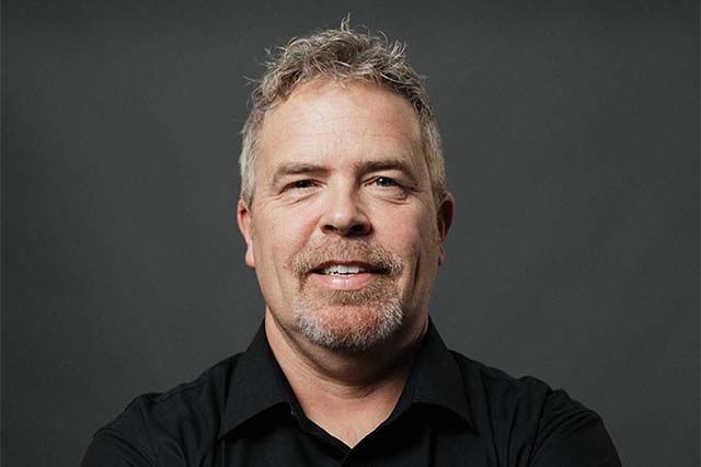 Man in navy polo shirt smiles, stands near brick wall.