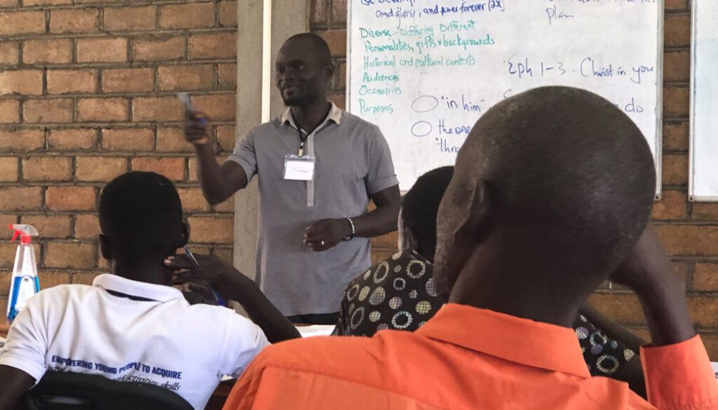 Man in grey shirt presenting to a group in a classroom setting, using a marker. Whiteboard visible.