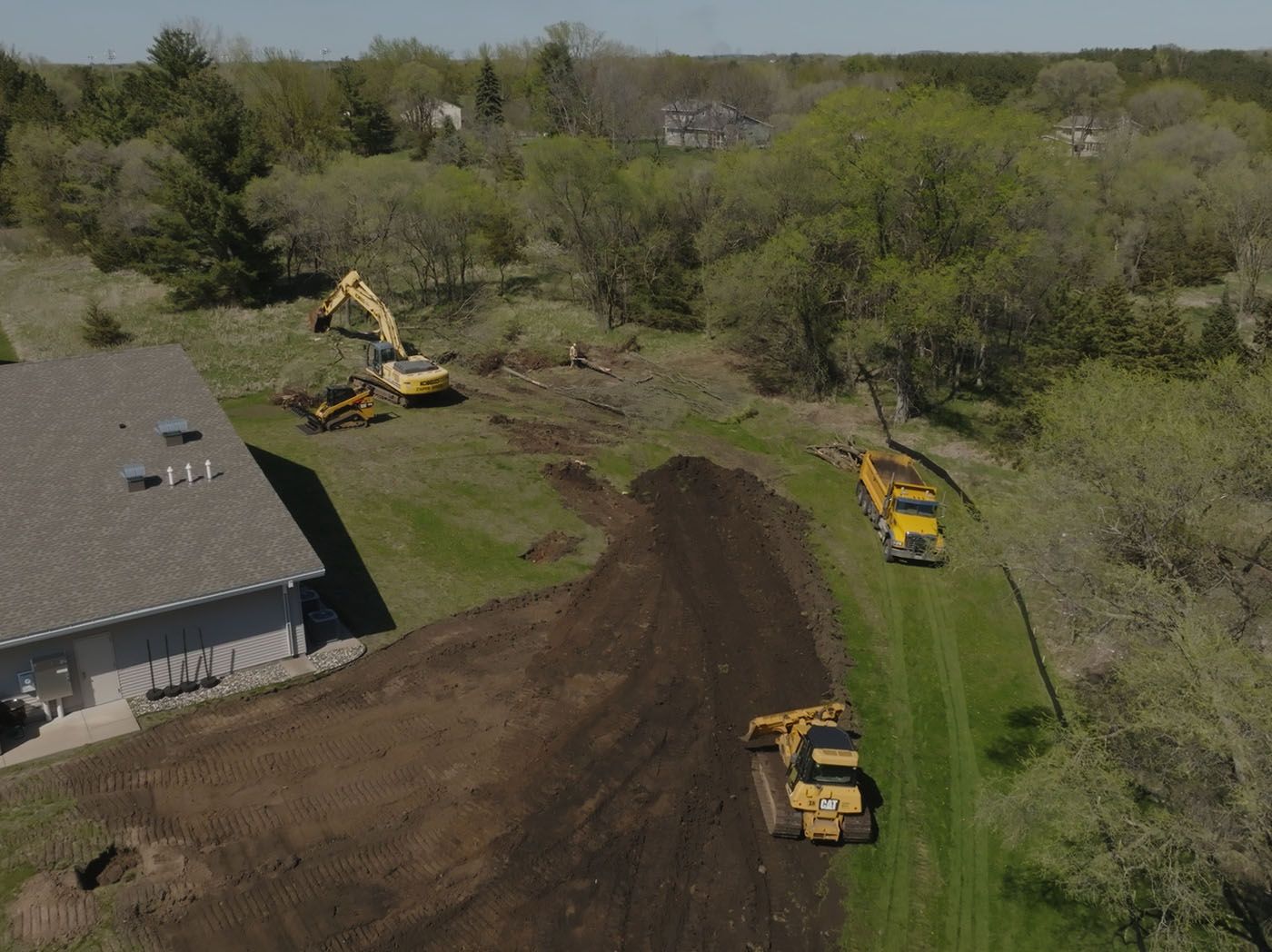 Construction site with excavators and dump trucks grading land near a house and trees.
