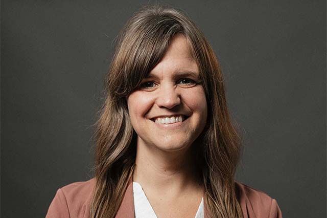 Woman smiling, wearing a denim shirt, outside in front of a building with glass doors.