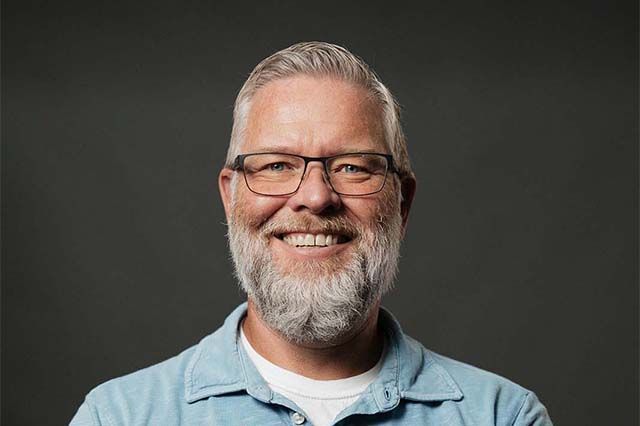 Man with beard smiles, arms crossed; blue shirt, brick building background.