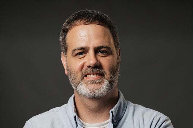 Man with a beard smiling, wearing a blue shirt. Outdoors, soft lighting.