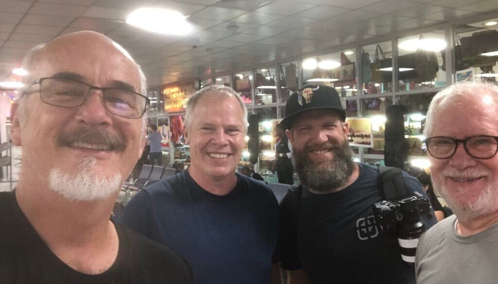 Four men smiling together inside a building with a brightly lit background.