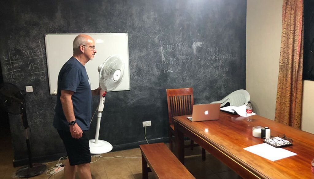 Man standing near a fan, pointing it toward himself. Room with a whiteboard and long wooden table.