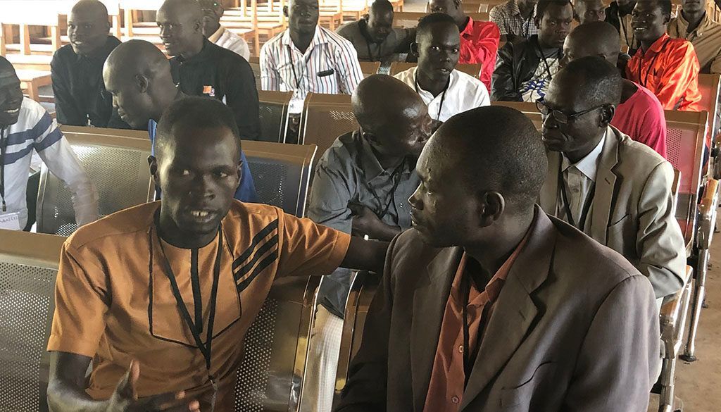 People sitting in rows, attending a meeting. Some are talking, wearing various colored shirts and suits.