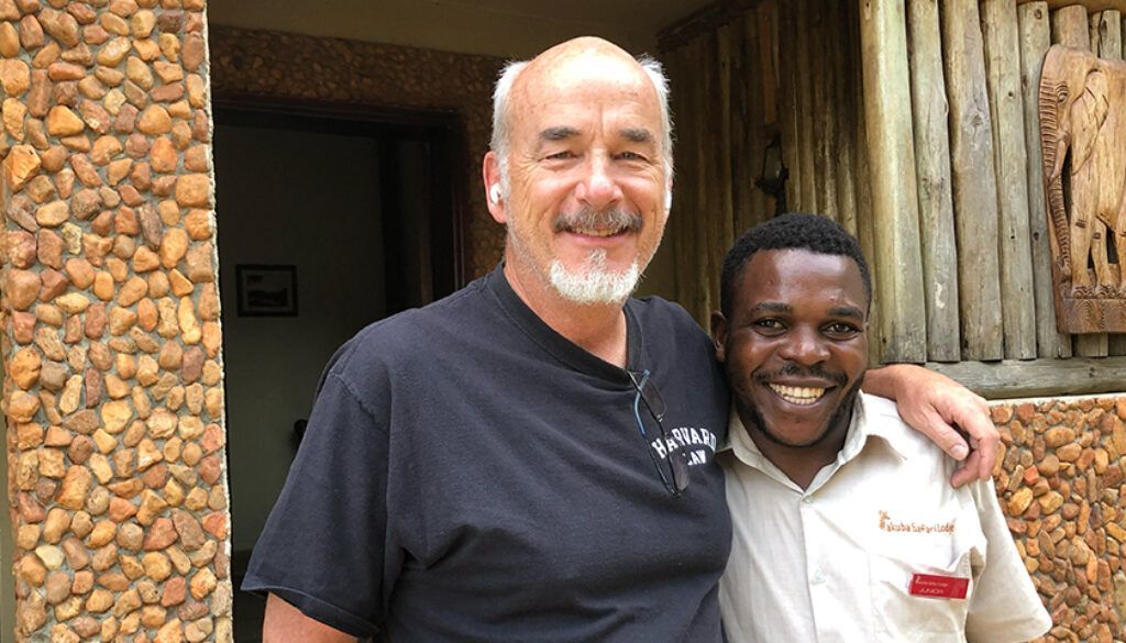 Man in black shirt with arm around smiling man in tan shirt by a stone wall.
