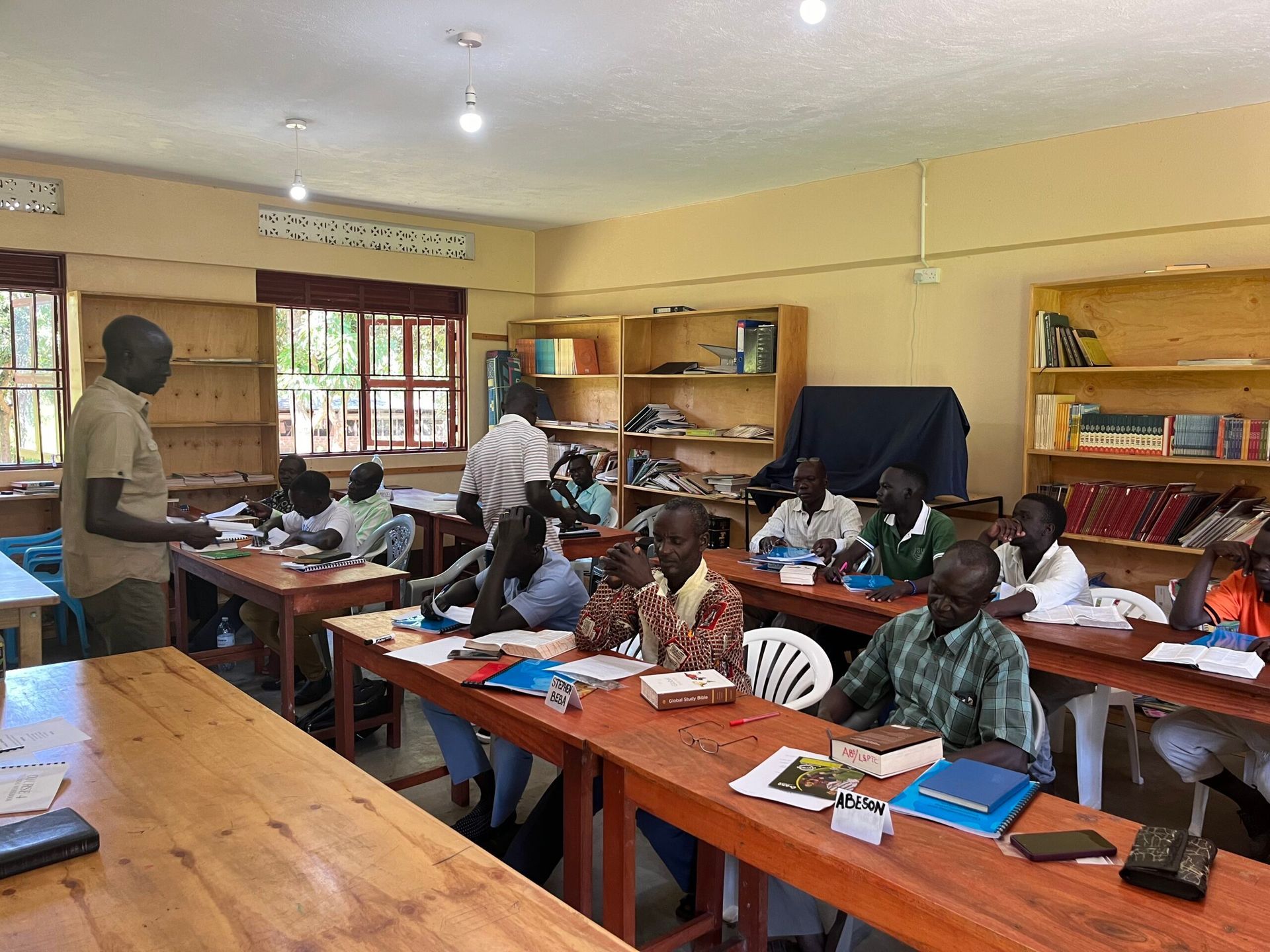 Classroom with students and a teacher. Students seated at desks, books and papers. Teacher stands, addressing them.