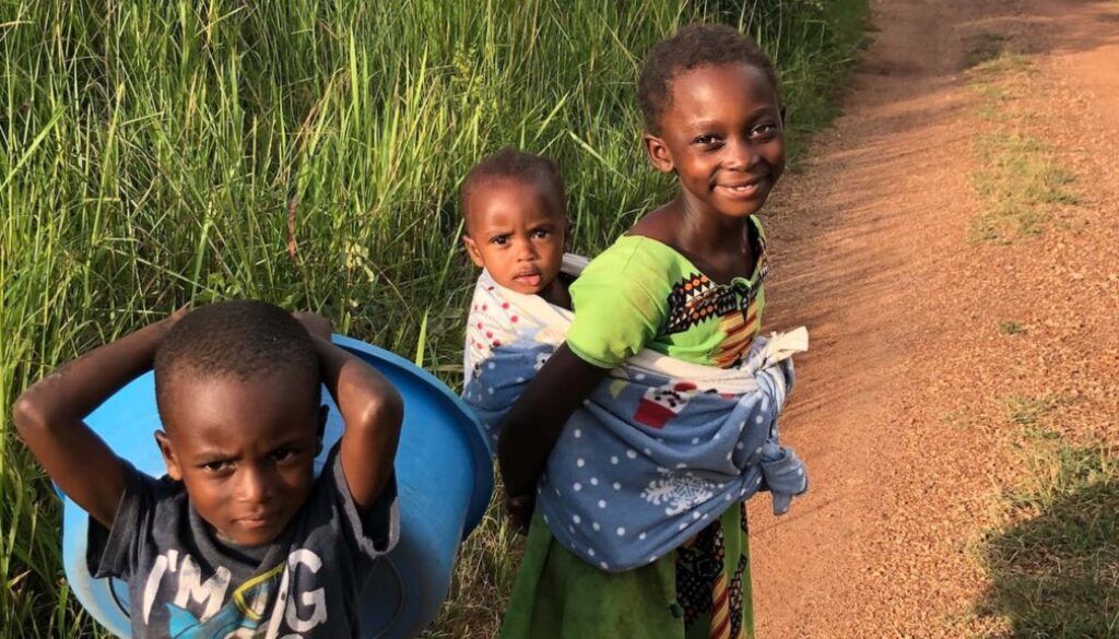 Three children on a dirt path: one carries a bucket, one is carried, the other smiles. Green grass, sunlit.