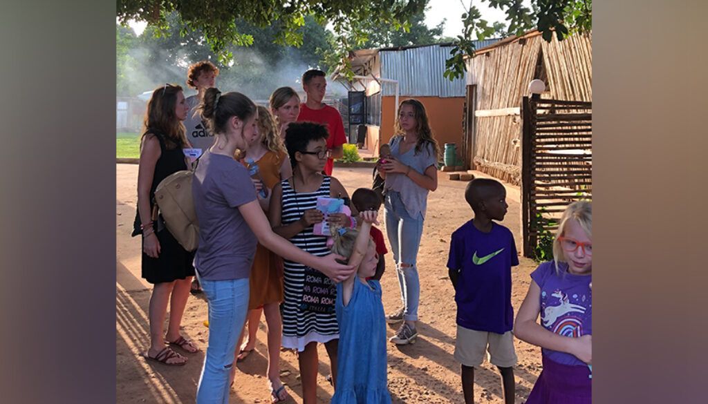 Group of people gathered outdoors, some interacting. Setting appears to be a village with simple buildings.