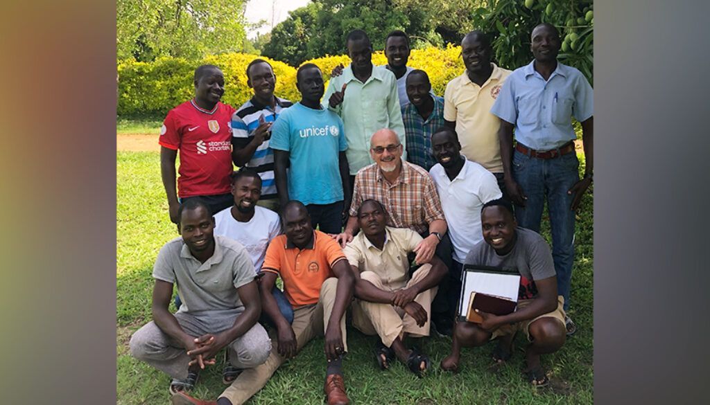 Group of men in a grassy area, some standing, some kneeling. One man with a beard sits in the middle.