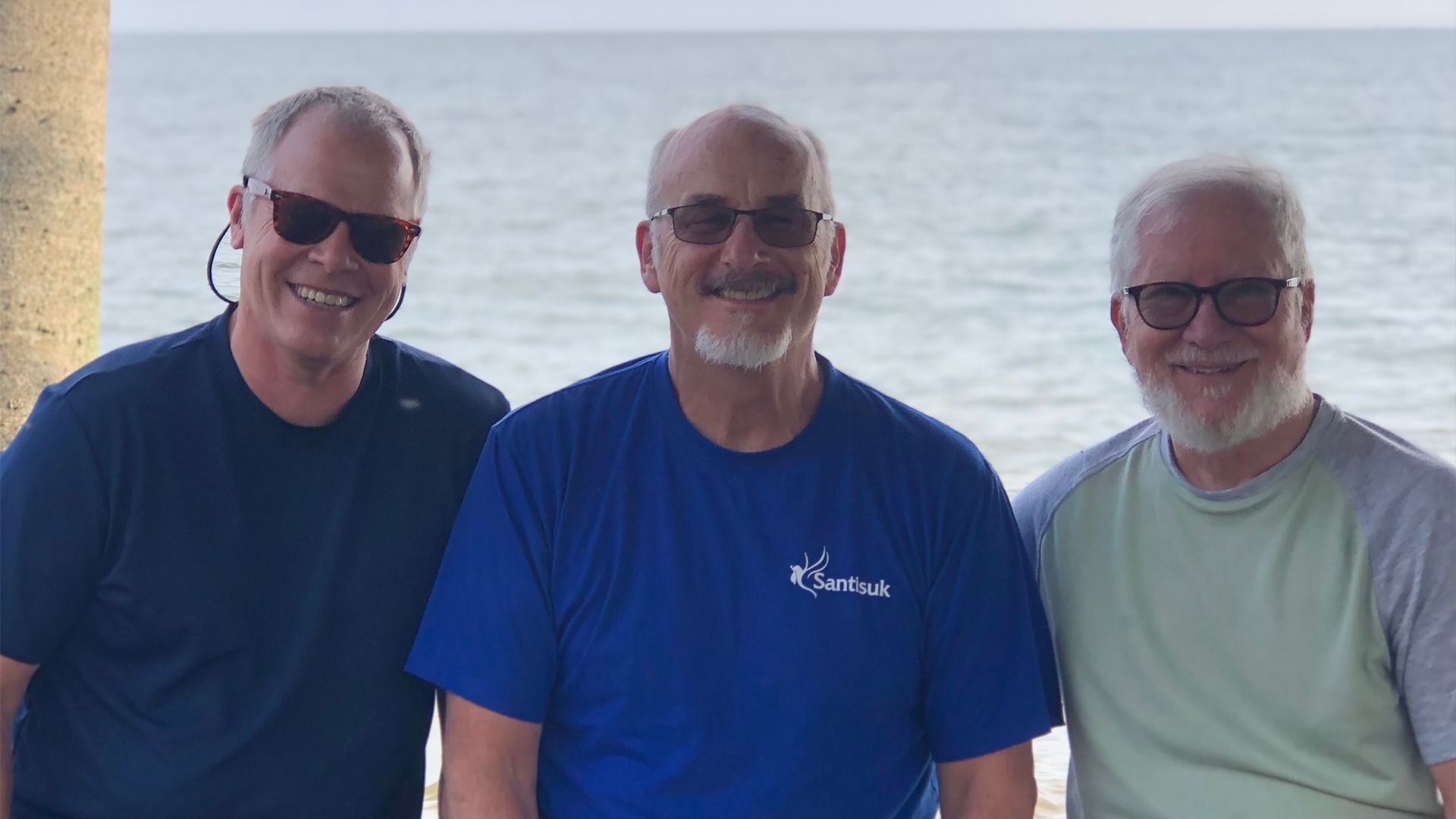Three men smiling in front of the ocean. They are wearing sunglasses and casual shirts.