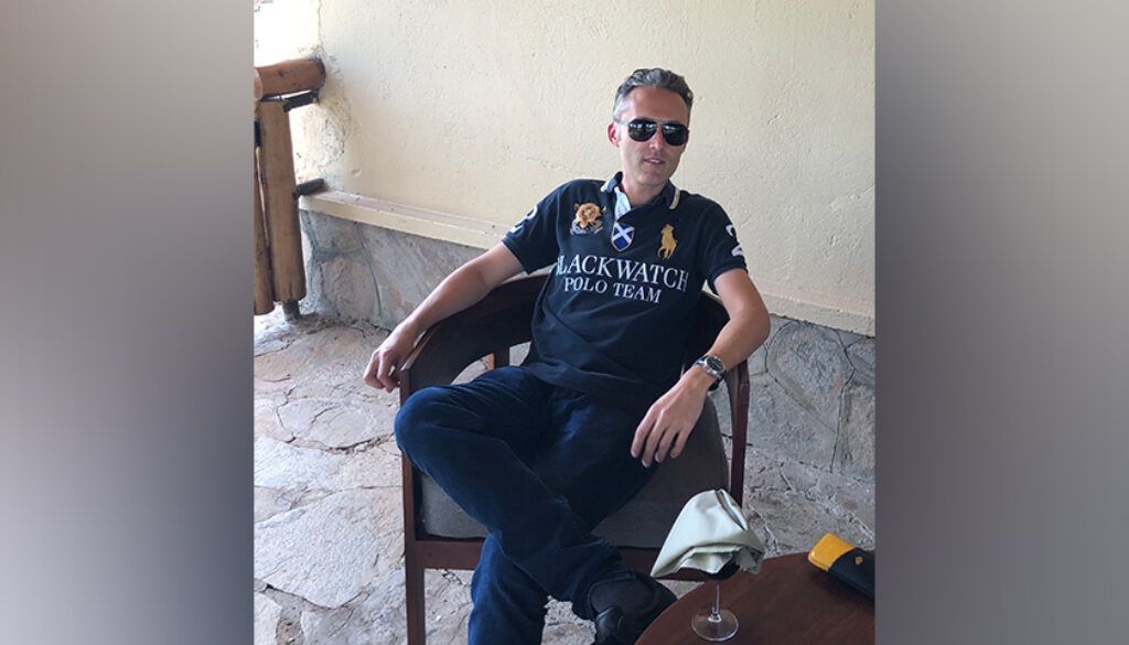 Man in sunglasses sits in a chair, wearing a dark polo shirt and jeans. Outdoors, with a table nearby.