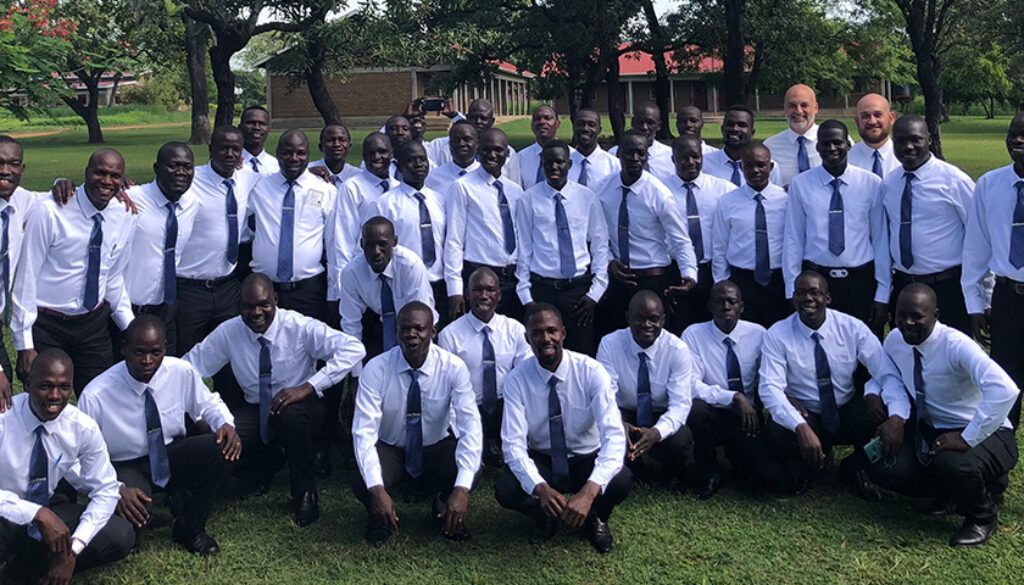 Group of men, wearing white shirts and ties, pose outdoors. Some squat, others stand. Green grass and trees visible.