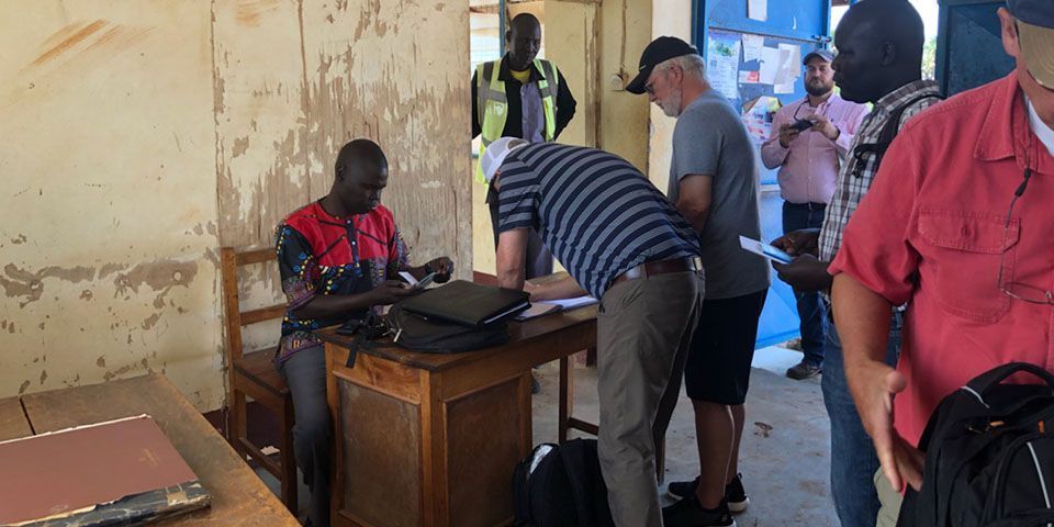 People registering at a desk in a room. A man in a striped shirt leans over the desk. Others stand nearby.