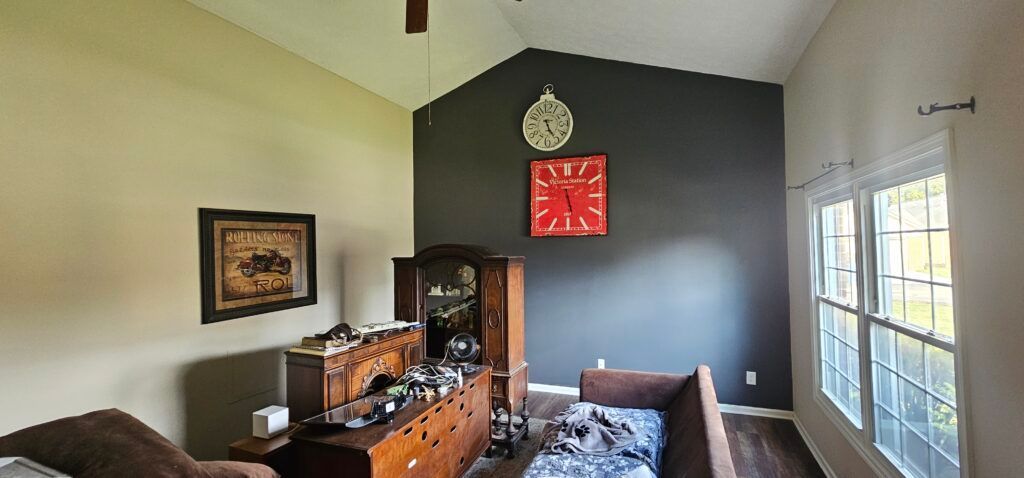 A room with a dark accent wall, furniture, clocks, a window with curtains, and a ceiling fan.