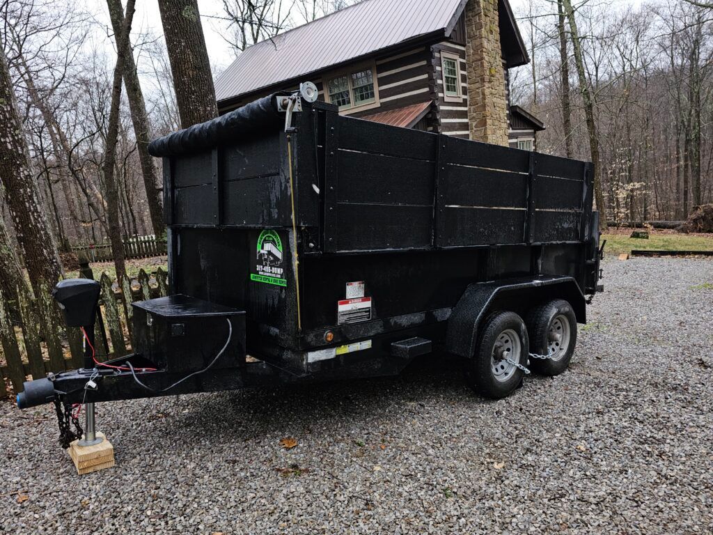 Black dump trailer parked on gravel, with a log cabin in the background.