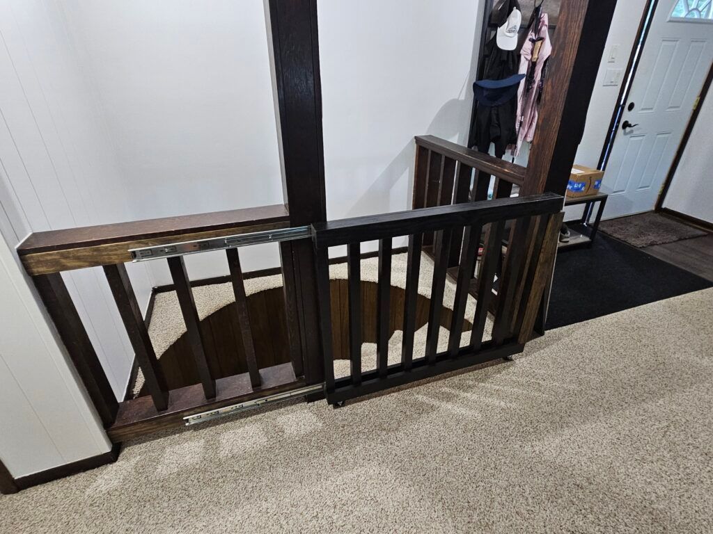 Dark brown baby gate at top of staircase, protecting an opening to the stairs. Beige carpet.