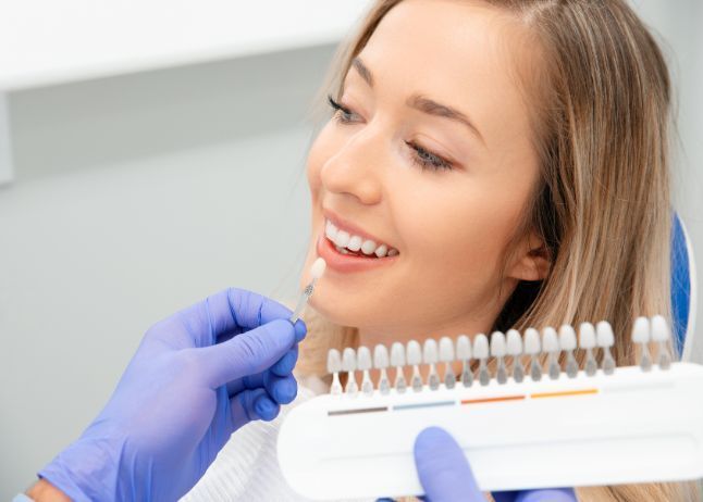 Dentist matching a woman's tooth shade using a color guide in a dental office setting.